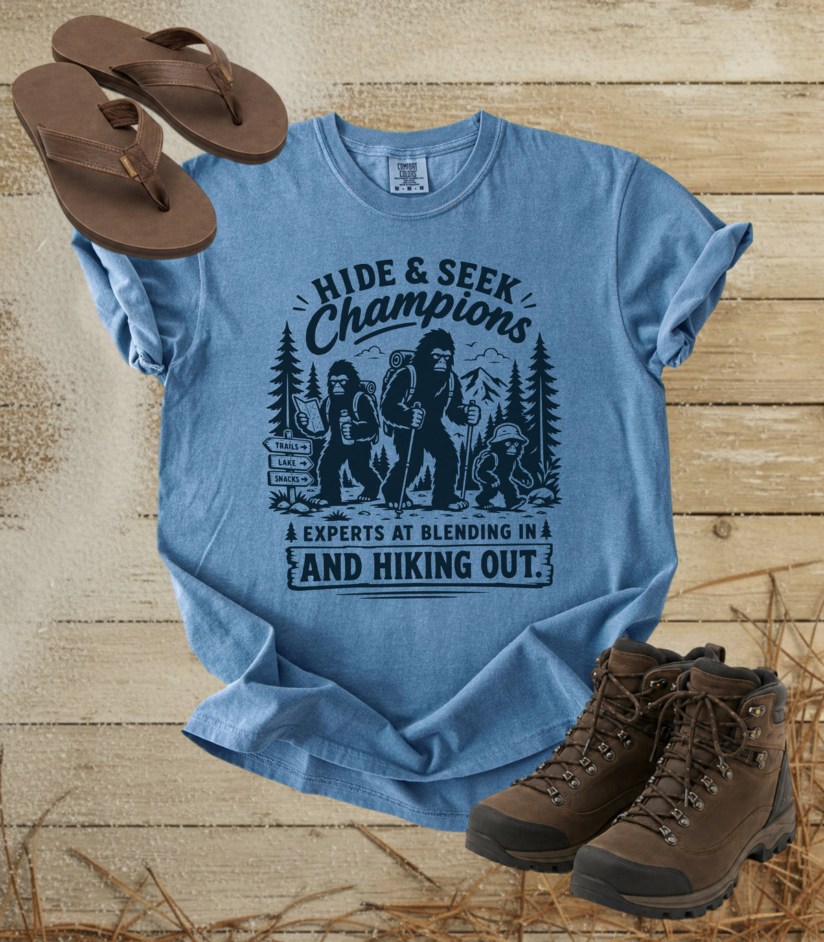 A comfort colors bluejean color graphic t-shirt laid flat on a rustic wood background, styled with brown flip-flops and hiking boots. The t-shirt features a dark blue graphic of a Bigfoot and other wild animals with the text 'HIDE & SEEK Champions' and 'A EXPERTS AT BLENDING IN A AND HIKING OUT.