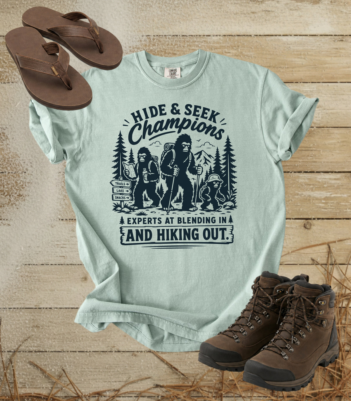 A comfort colors bay color graphic t-shirt featuring a black print of a Bigfoot and hiking buddies with the text 'HIDE & SEEK Champions'. The shirt is styled in a flat-lay on a rustic wooden background, accompanied by brown flip-flops and hiking boots.