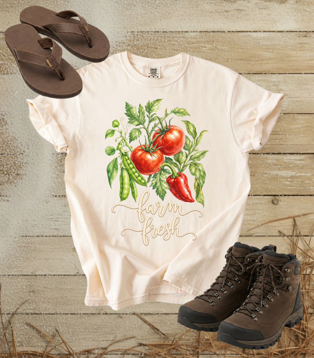 A comfort colors ivory color short-sleeve t-shirt with a watercolor graphic of tomatoes, green peppers, and peas, with the text 'Harvest Fresh' written below. The shirt is styled in a flat-lay arrangement on a rustic wooden background, accompanied by brown leather sandals and hiking boots.