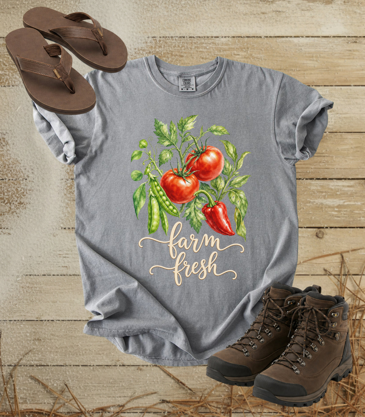A comfort colors grey color graphic T-shirt laid flat on a rustic wood surface, featuring a watercolor-style tomato and pepper illustration with the words “farm fresh.” Brown sandals are placed near the top left and brown hiking boots near the bottom right as styling props.