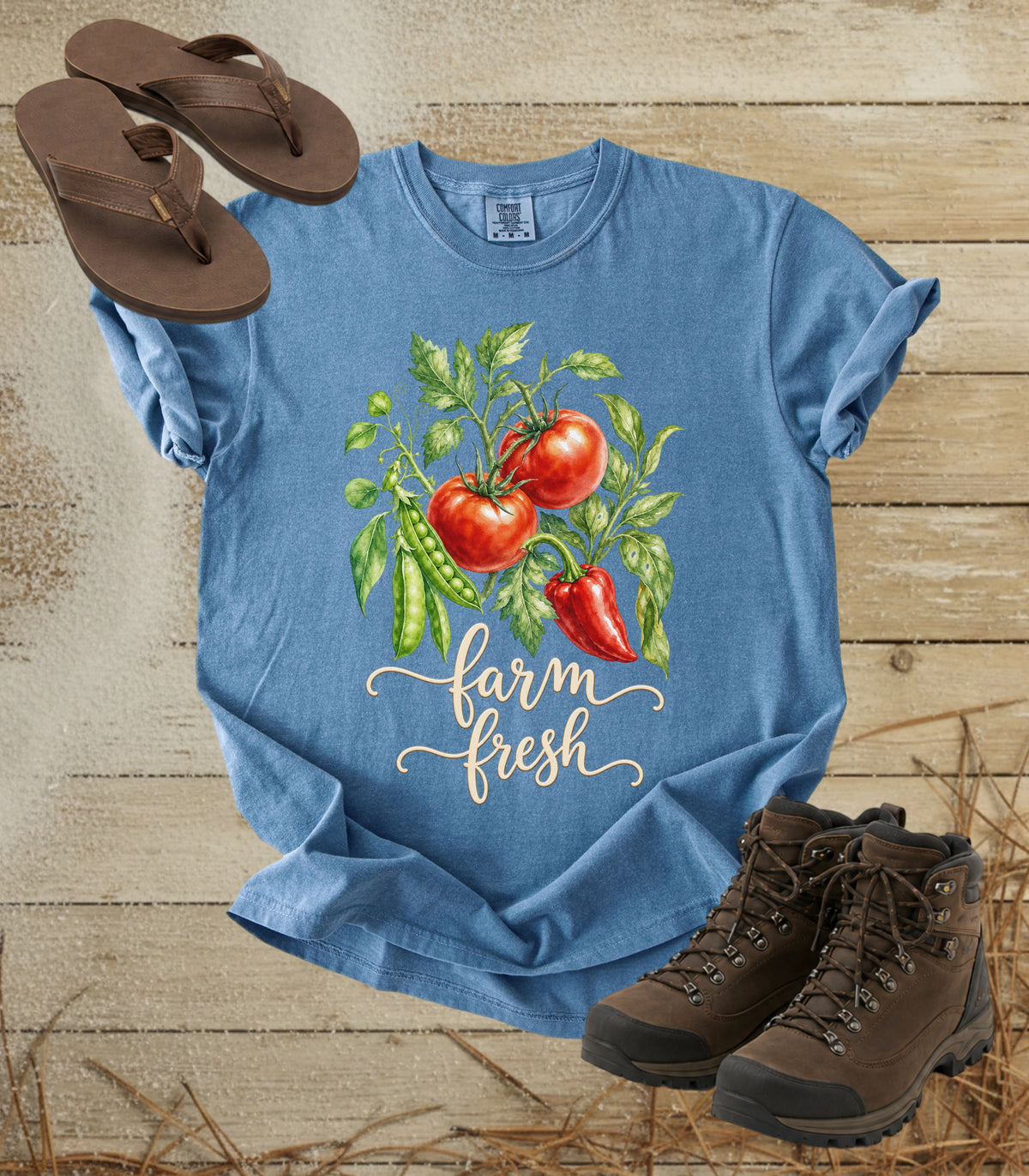 A comfort colors bluejean color short-sleeve graphic T-shirt laid flat on a rustic wood surface, styled with brown flip-flops and brown hiking boots. The shirt features a watercolor-style tomato plant graphic with red tomatoes, green leaves, and the words 'farm fresh' in white script.