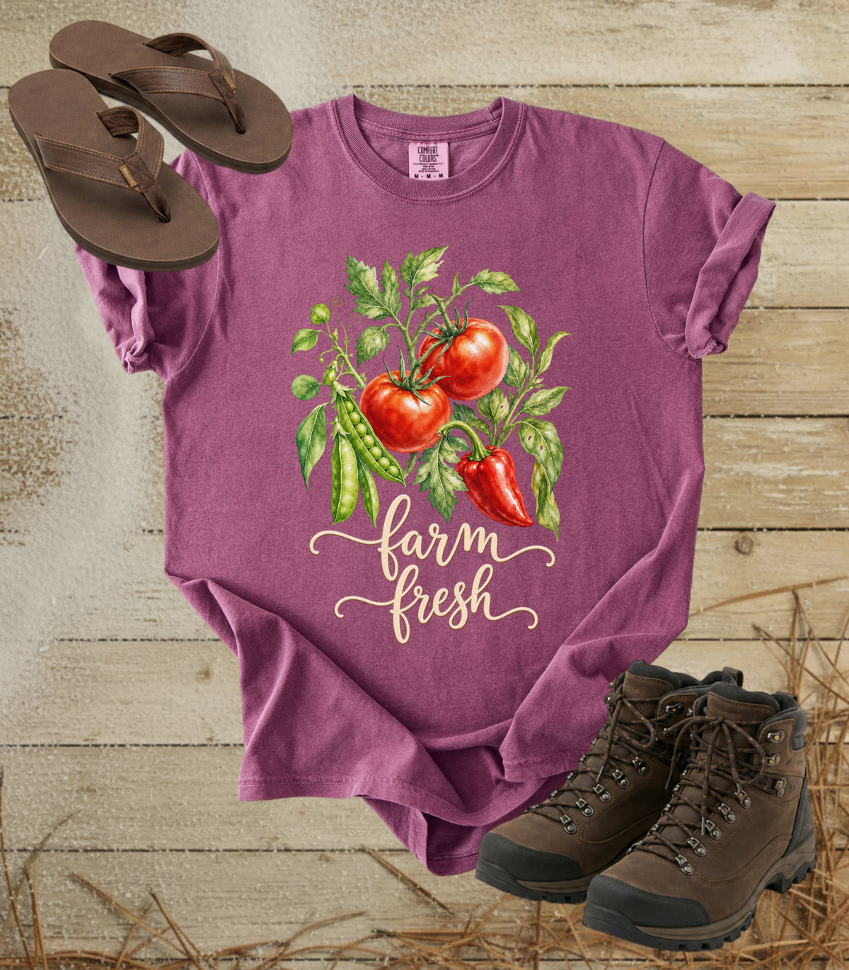 A comfort colors berry color short-sleeve t-shirt with a 'farm fresh' graphic featuring tomatoes, green peas, and a red pepper, styled in a flat lay with brown sandals and hiking boots on a rustic wood background.