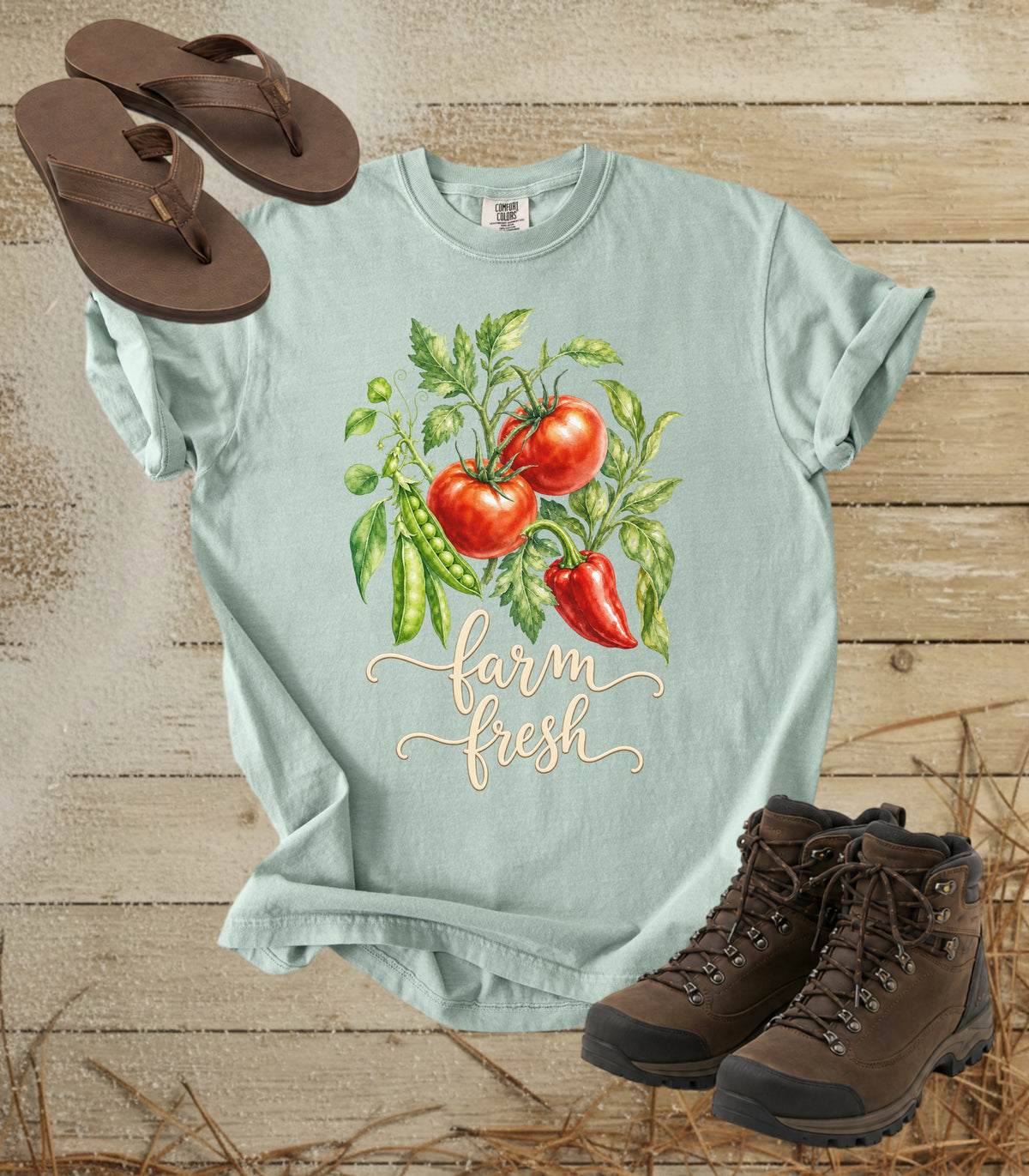 A comfort colors bay color crewneck t-shirt with a watercolor graphic of tomatoes, green peas, and a red pepper, with the words 'farm fresh' in script. The shirt is styled in a flat lay on a rustic wood background with brown flip-flops and hiking boots.