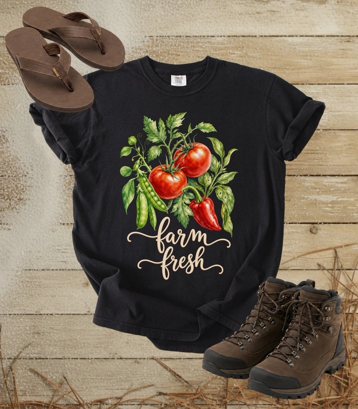 A comfort colors black color graphic t-shirt featuring a watercolor illustration of red tomatoes, green peas, and a red pepper with the text 'farm fresh' in white script. The shirt is styled in a flat-lay arrangement on a rustic wooden background, accompanied by brown flip-flops and hiking boots.