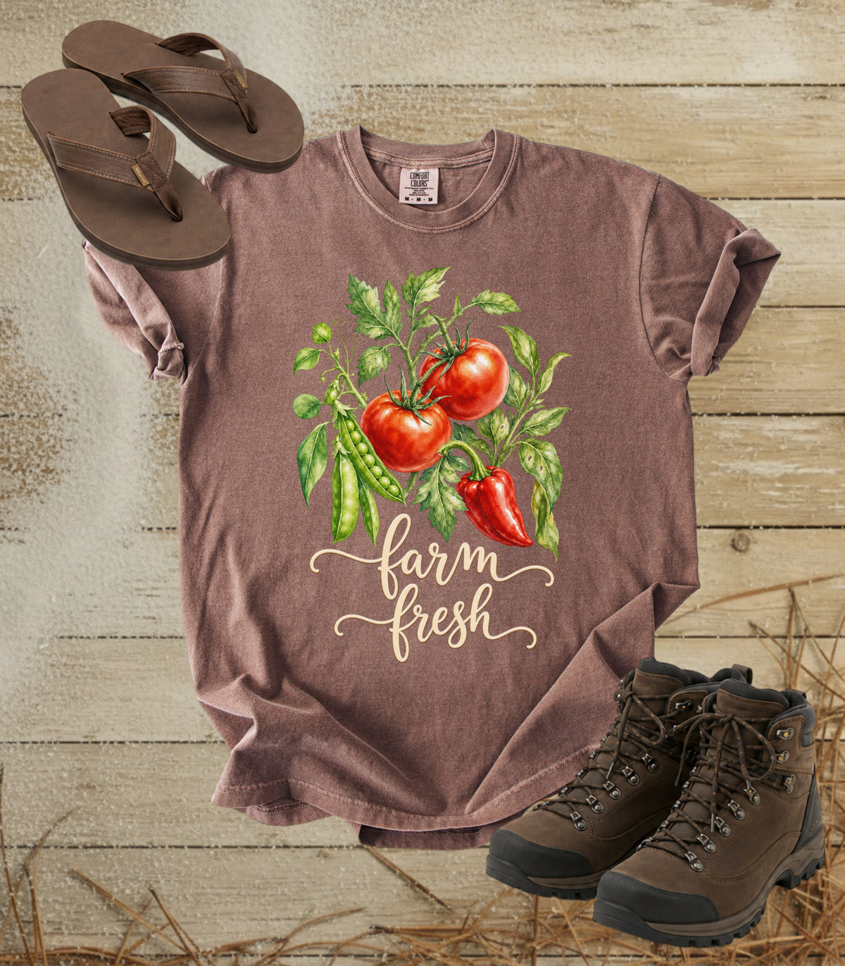 A comfort colors espresso color graphic t-shirt laid flat on a rustic wood background, styled with brown flip-flops and hiking boots. The t-shirt features a watercolor graphic of red tomatoes, green peas, a red chili pepper, and the words 'farm fresh' in beige script.