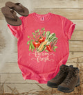 A comfort colors watermelon color short-sleeve graphic T-shirt laid flat on a rustic wood plank surface, styled with brown flip-flops, brown hiking boots, and straw. The shirt features a colorful vegetable illustration with the words “farm fresh” in white script.