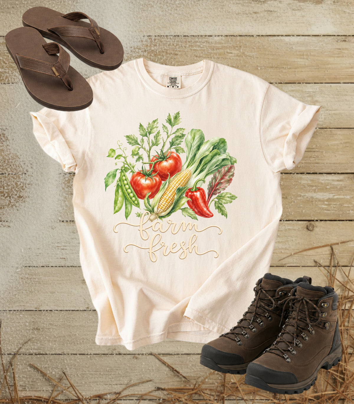A comfort colors ivory color short-sleeve t-shirt with a colorful watercolor vegetable graphic and the words 'farm fresh' in gold script, styled in a flat lay with brown sandals, hiking boots, and straw on a rustic wood plank background.