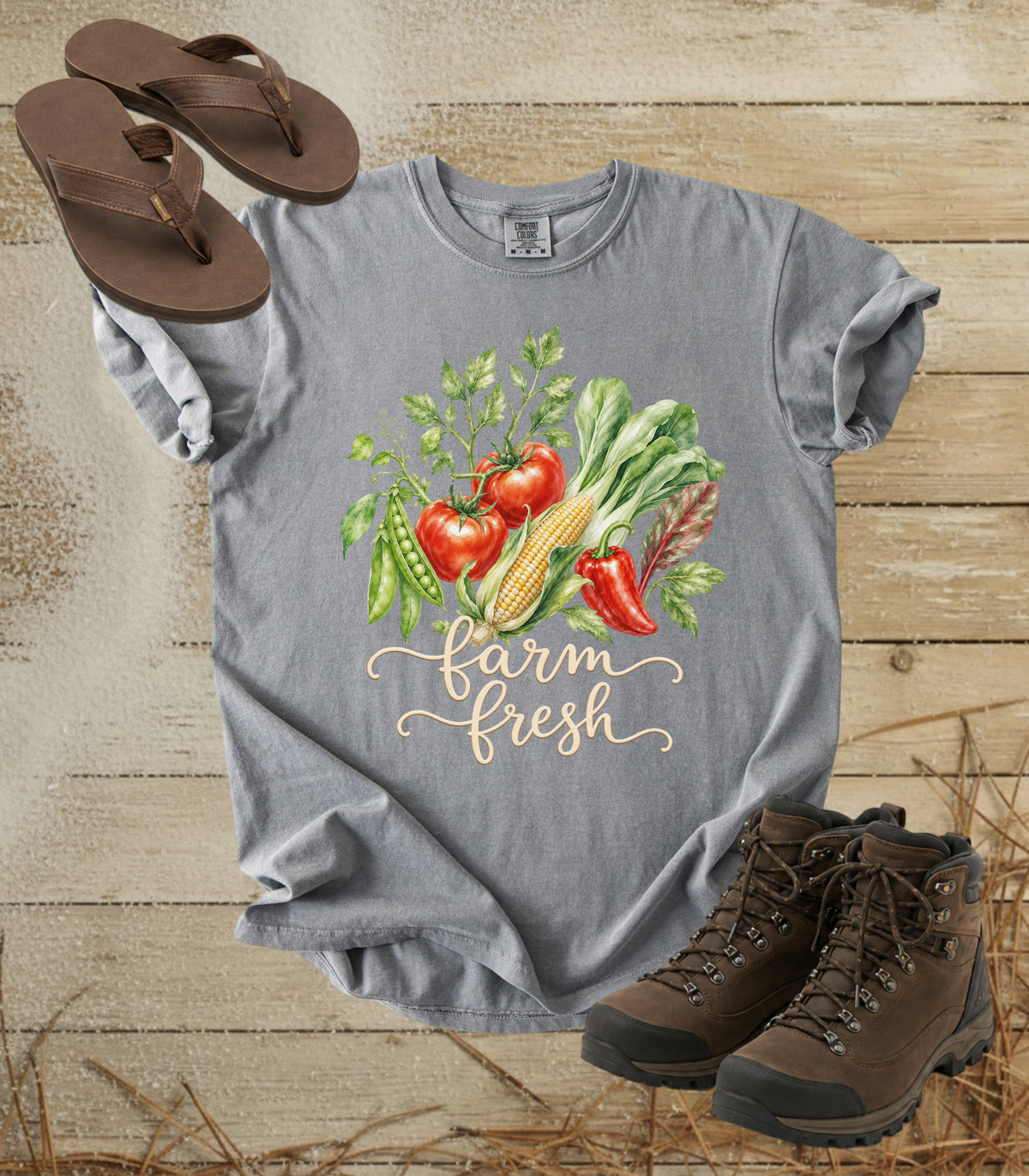 A comfort colors grey color graphic T-shirt laid flat on a rustic wood plank surface, styled with brown flip-flops and brown hiking boots. The shirt features a colorful watercolor-style vegetable illustration with tomatoes, corn, peas, lettuce, and red peppers above the words “farm fresh” in a light beige script.