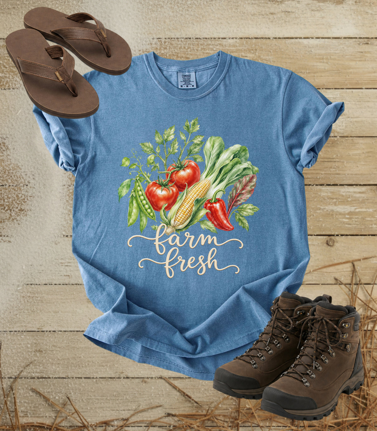 A comfort colors bluejean color short-sleeve T-shirt laid flat on a rustic wood surface, printed with a colorful vegetable illustration and the words 'farm fresh.' Brown sandals are placed near the top left, and brown hiking boots are placed near the bottom right.