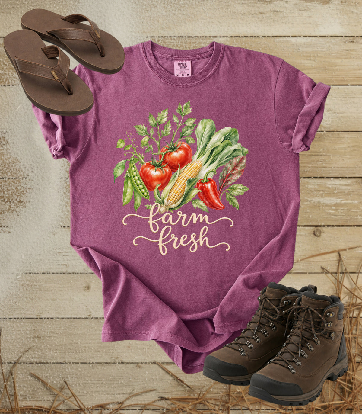 A comfort colors berry color short-sleeve t-shirt with a colorful graphic of fresh vegetables and the text 'farm fresh' in script. The shirt is styled in a flat lay on a rustic wooden background, accompanied by brown flip-flops and hiking boots.