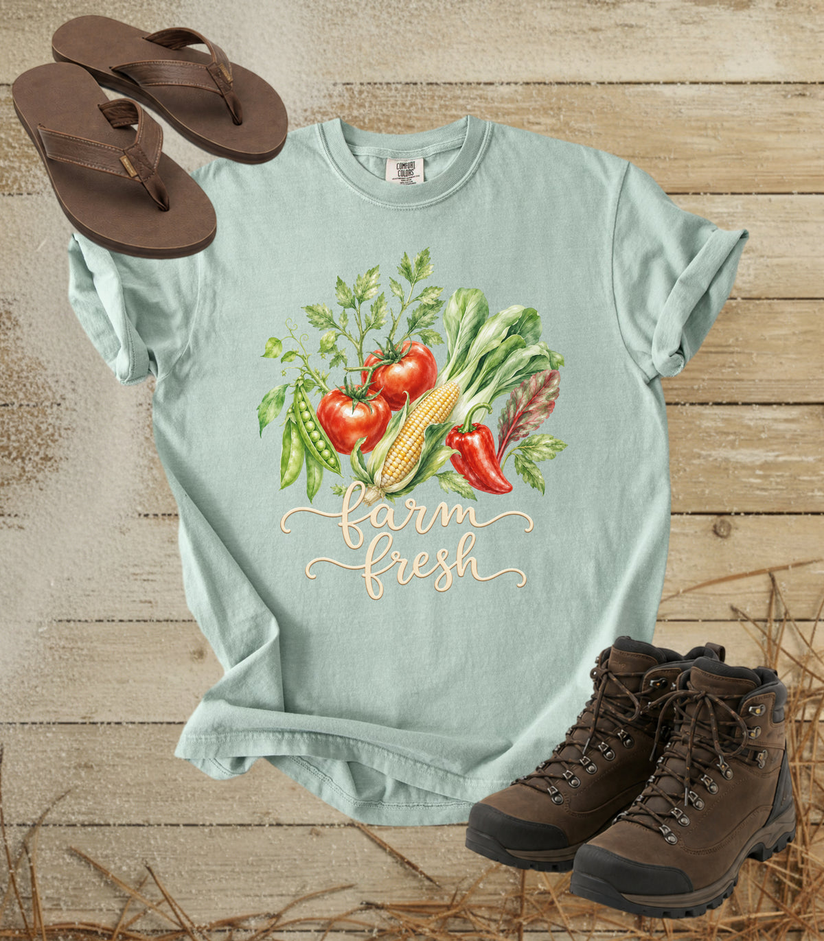 A comfort colors bay color graphic t-shirt with a 'farm fresh' vegetable print, styled in a flat lay with brown flip-flops and hiking boots on a rustic wood background.