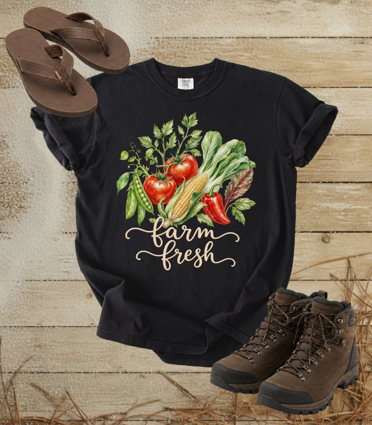 A comfort colors black color graphic t-shirt featuring a watercolor illustration of fresh farm vegetables (tomatoes, corn, peppers, greens) and the text 'farm fresh' in white script. The shirt is styled in a flat-lay arrangement with brown flip-flops and hiking boots on a rustic wooden background with straw.