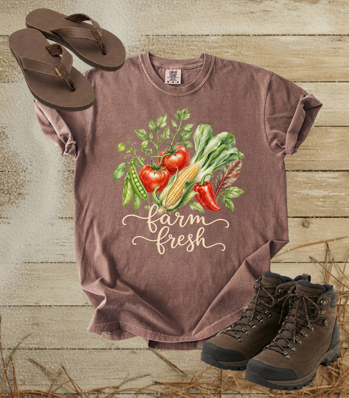 A comfort colors espresso color short-sleeve graphic T-shirt laid flat on a rustic wood plank background, styled with brown flip-flops and hiking boots. The shirt features a colorful watercolor-style vegetable print with the words 'farm fresh' in script.