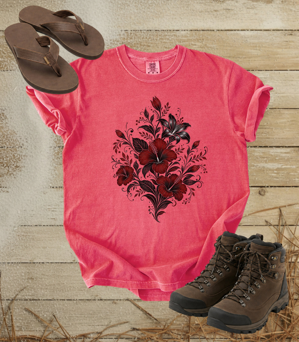 A comfort colors watermelon color short-sleeve t-shirt with a black and red floral graphic on the front, styled in a flat lay with brown flip-flops, brown hiking boots, and straw on a rustic wooden background.