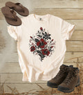 A comfort colors ivory color short-sleeve t-shirt with a red and black floral heart graphic, styled in a flat lay with brown leather sandals and hiking boots on a rustic wood plank background.