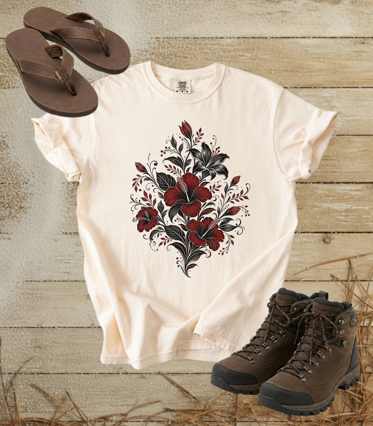 A comfort colors ivory color short-sleeve t-shirt with a red and black floral heart graphic, styled in a flat lay with brown leather sandals and hiking boots on a rustic wood plank background.