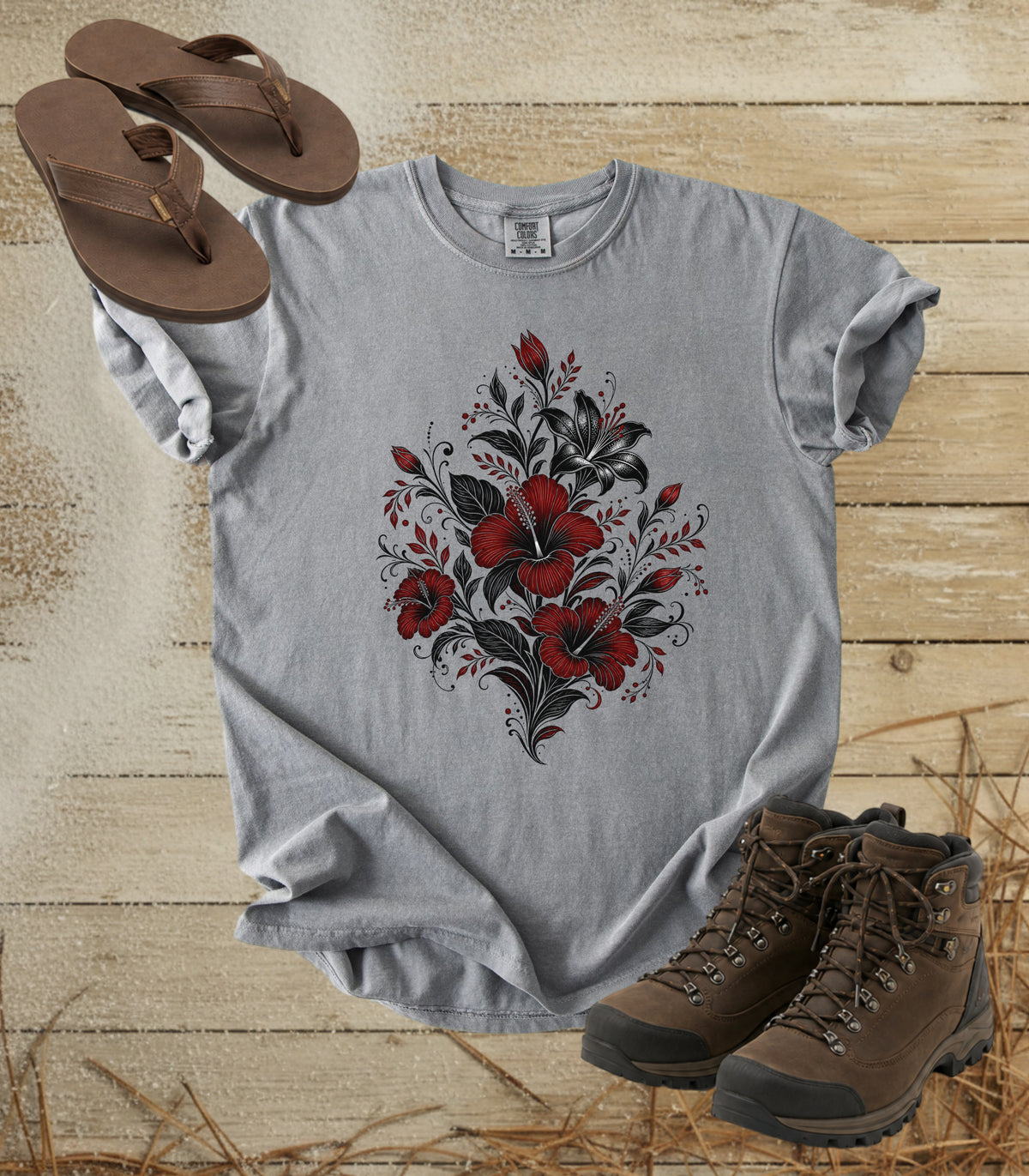 A comfort colors grey color short-sleeve t-shirt with a red and black floral heart graphic on the front, styled in a flat lay on a rustic wooden background with brown sandals and hiking boots.