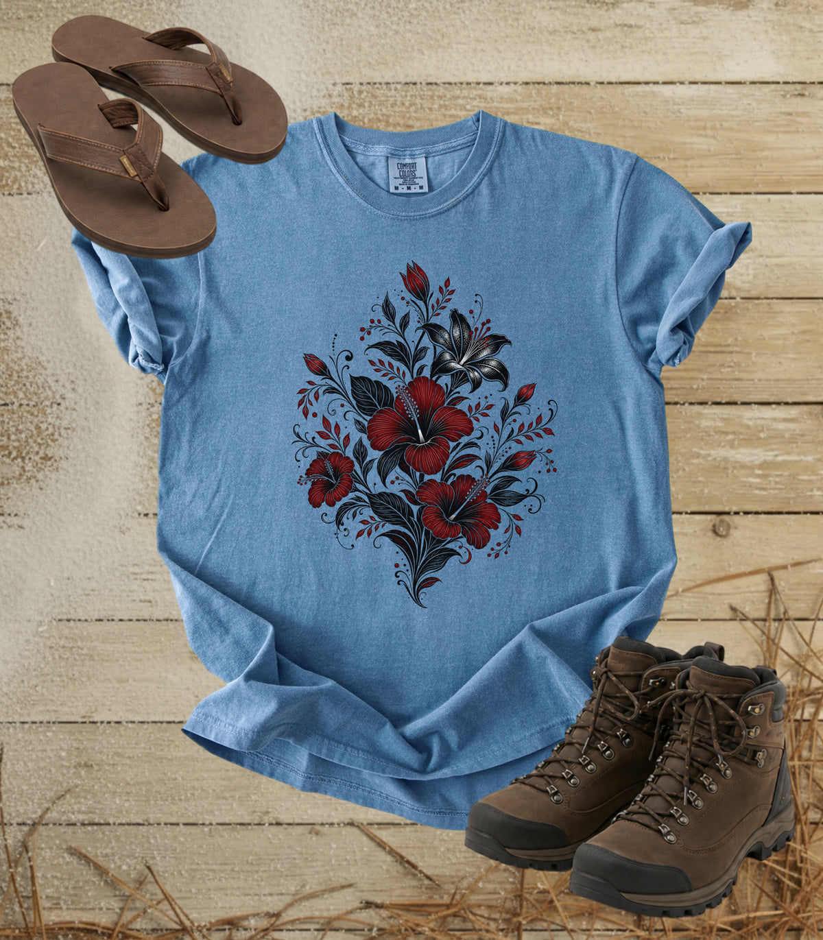 A comfort colors bluejean color t-shirt with a red and black floral graphic on the front, styled in a flat lay with brown flip-flops and hiking boots on a rustic wooden background.