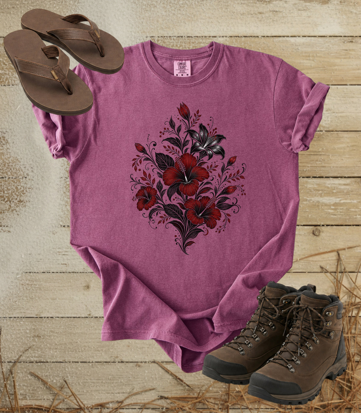 A comfort colors berry color short-sleeve t-shirt with a red and black floral graphic on the front, styled in a flat lay with brown flip-flops and hiking boots on a rustic wood plank background.