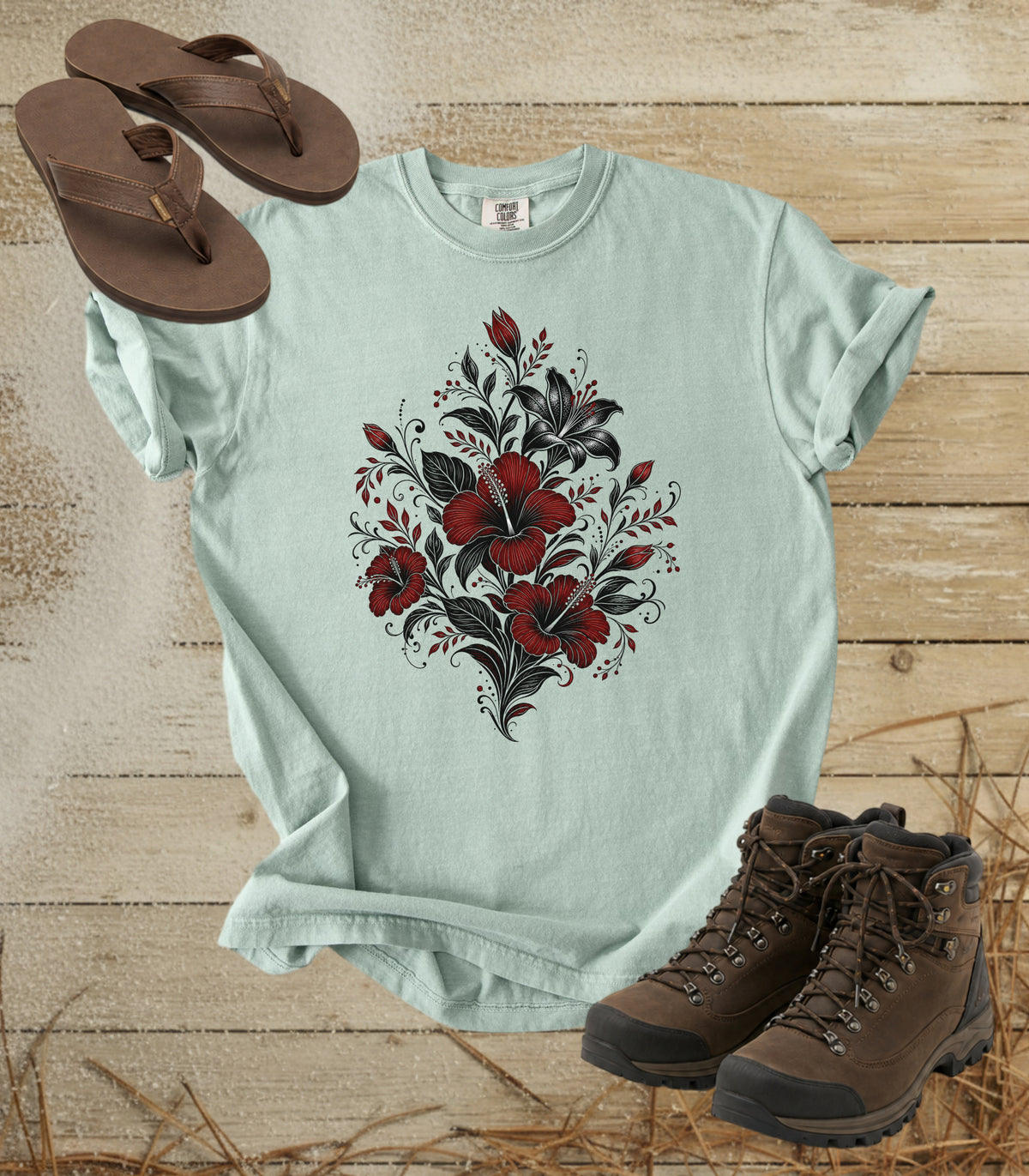 A comfort colors bay color t-shirt with a red and black floral graphic on the front, styled in a flat lay with brown flip-flops and brown hiking boots on a rustic wooden background.