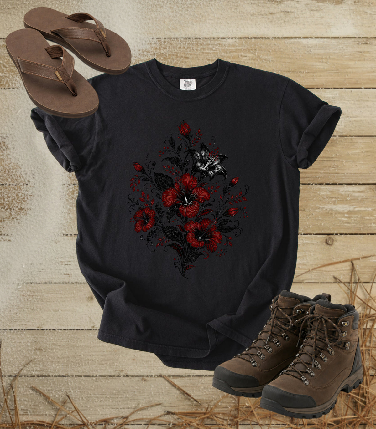 A comfort colors black color t-shirt with a red and black floral graphic on the front, styled in a flat-lay arrangement with brown flip-flops and brown hiking boots on a rustic wood plank background.