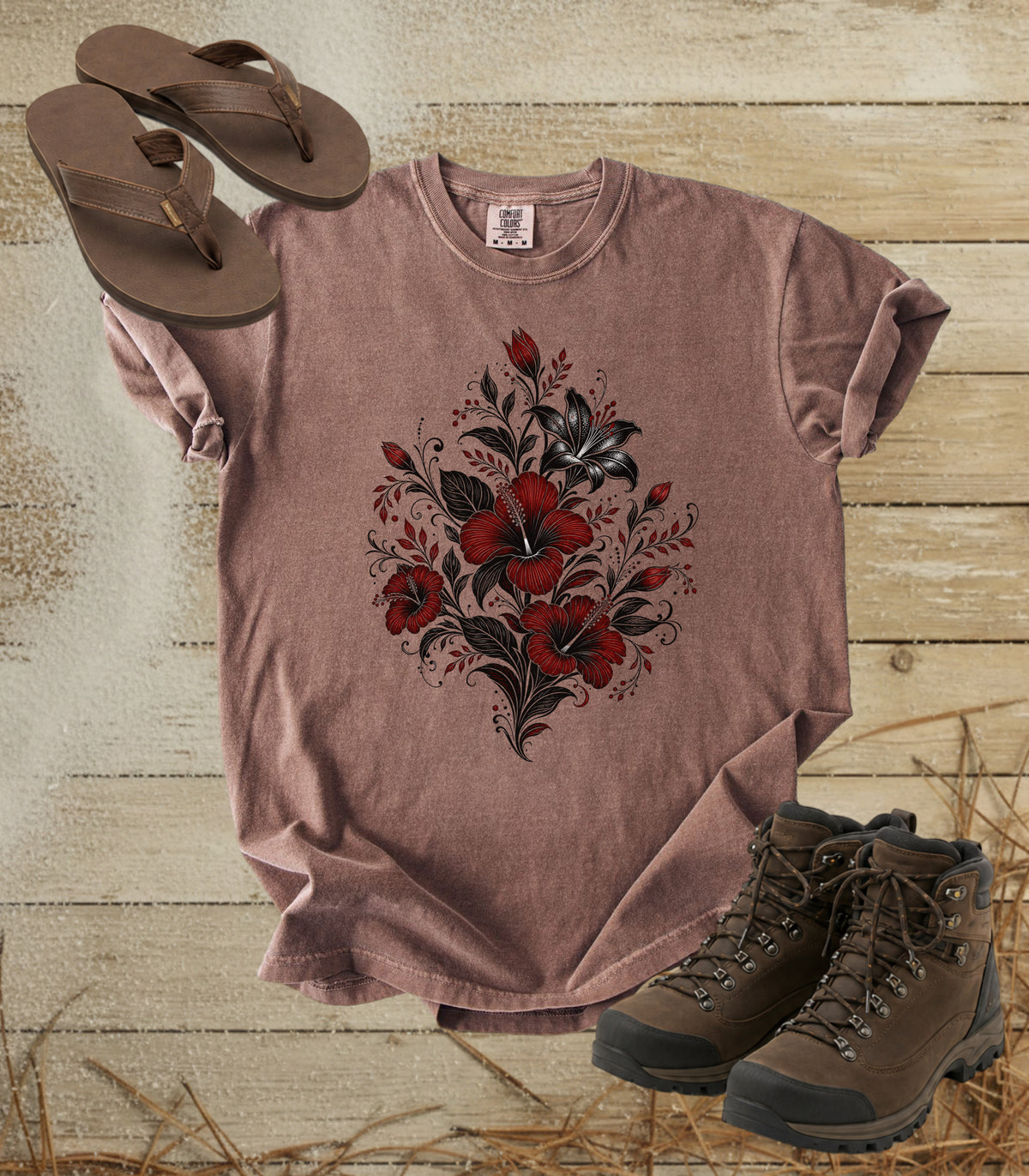 A comfort colors espresso color short-sleeve t-shirt with a red and black floral hibiscus print, styled in a flat-lay arrangement with brown flip-flops and hiking boots on a rustic wood plank background.